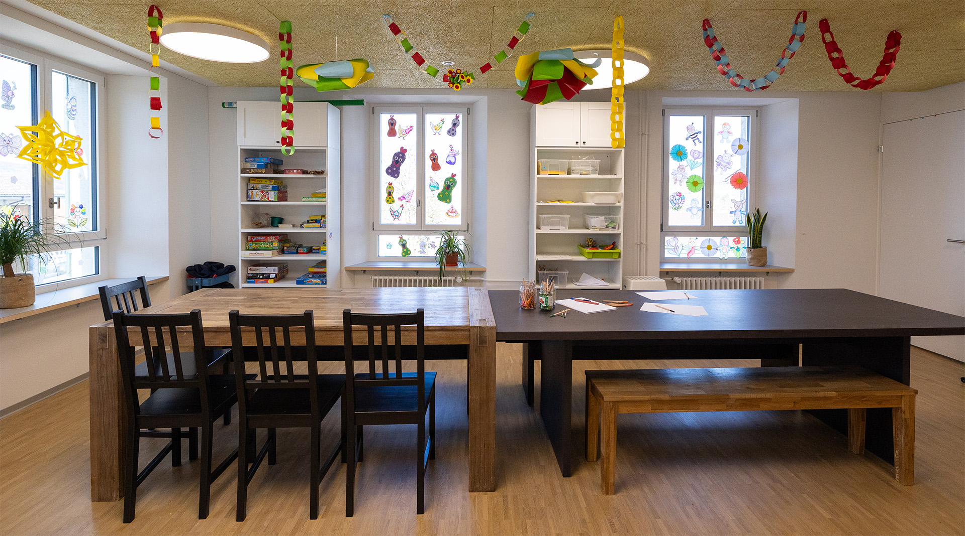 Freundlicher Gruppenraum der Tagesstrukturen Wohlen Halde mit Spielregalen, grossen Tischen und liebevoll dekorierten Fenstern.