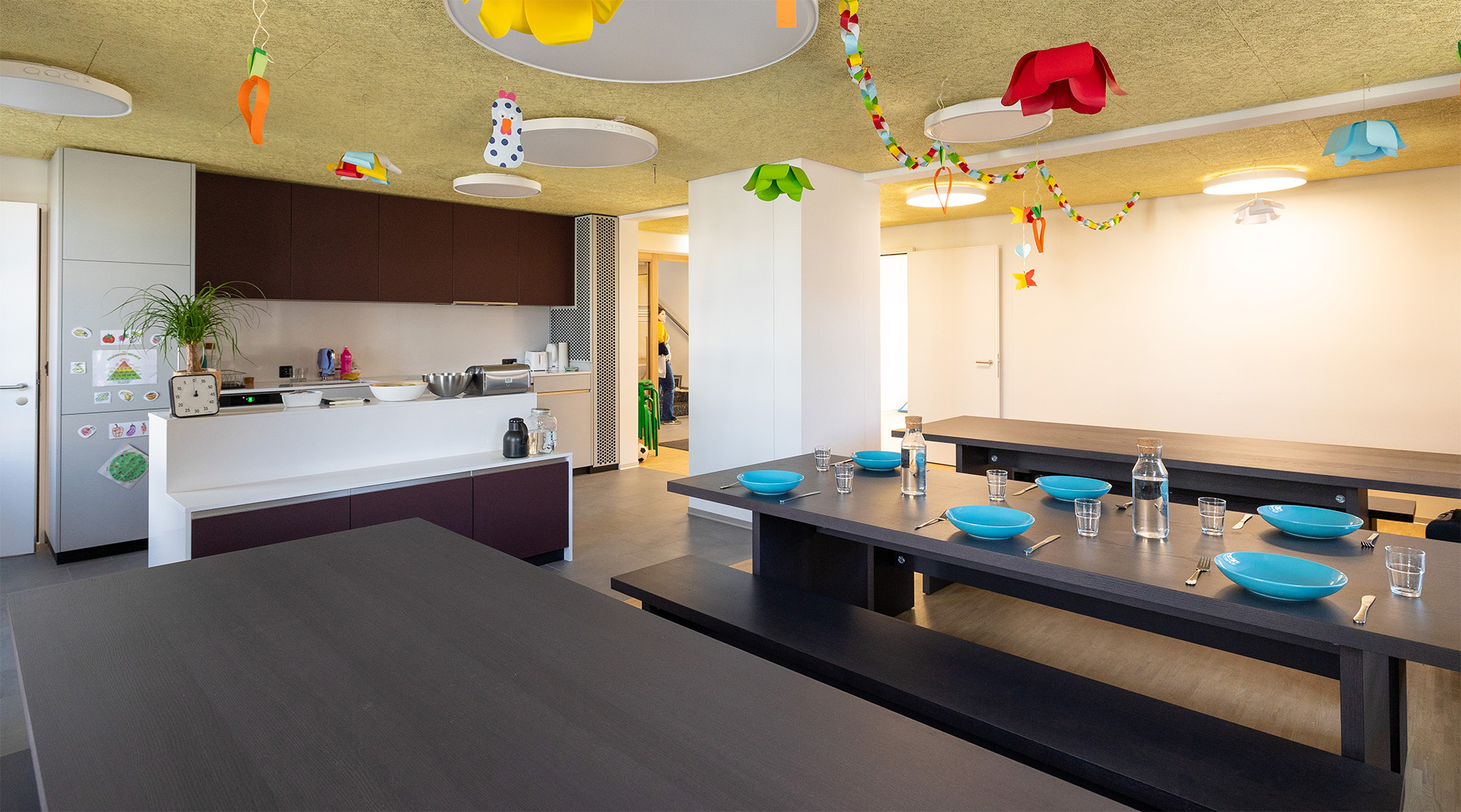 Heller Ess- und Küchenbereich der Tagesstrukturen Wohlen Halde mit langen dunklen Tischen, gedeckten Plätzen, moderner offener Küche und farbenfroher Kinderdekoration an der Decke.