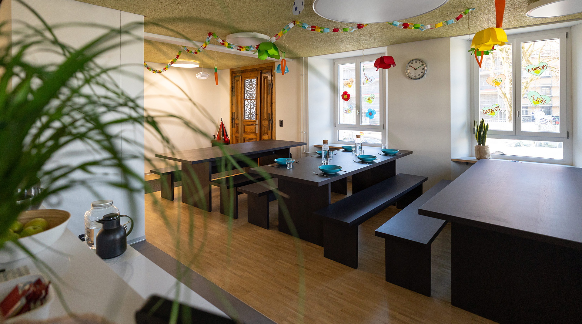 inladender Essbereich der Tagesstrukturen Wohlen Halde mit langen dunklen Tischen, Sitzbänken, gedeckten Plätzen, grossen Fenstern und farbenfroher Kinderdekoration.