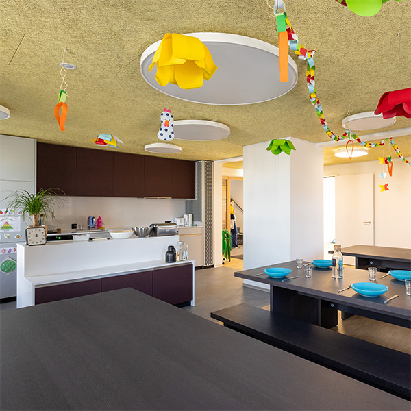 Heller Ess- und Küchenbereich der Tagesstrukturen Wohlen Halde mit langen dunklen Tischen, gedeckten Plätzen, moderner offener Küche und farbenfroher Kinderdekoration an der Decke.